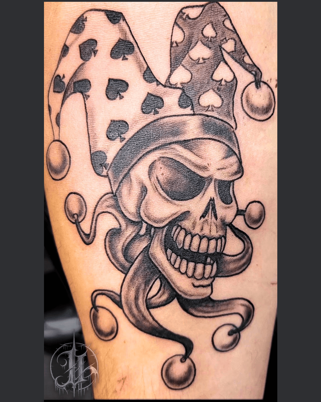 Black-and-grey jester skull tattoo with a spade-patterned hat and bells, on a lower leg.